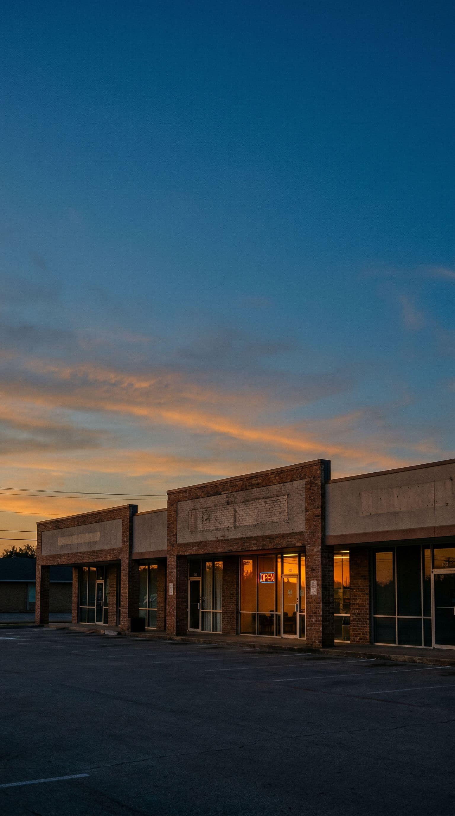 Texas commercial strip plaza at dusk, vertical composition with warm tenant lights and an OPEN sign glowing.