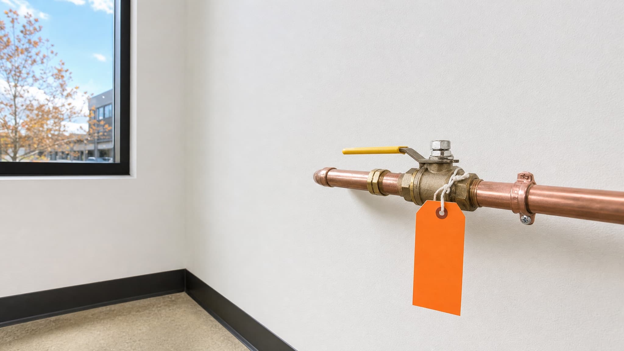 A brass ball shutoff valve with a yellow lever on a copper water line, with a blank orange tag tied to the handle, in a bright white utility room with an autumn window view.