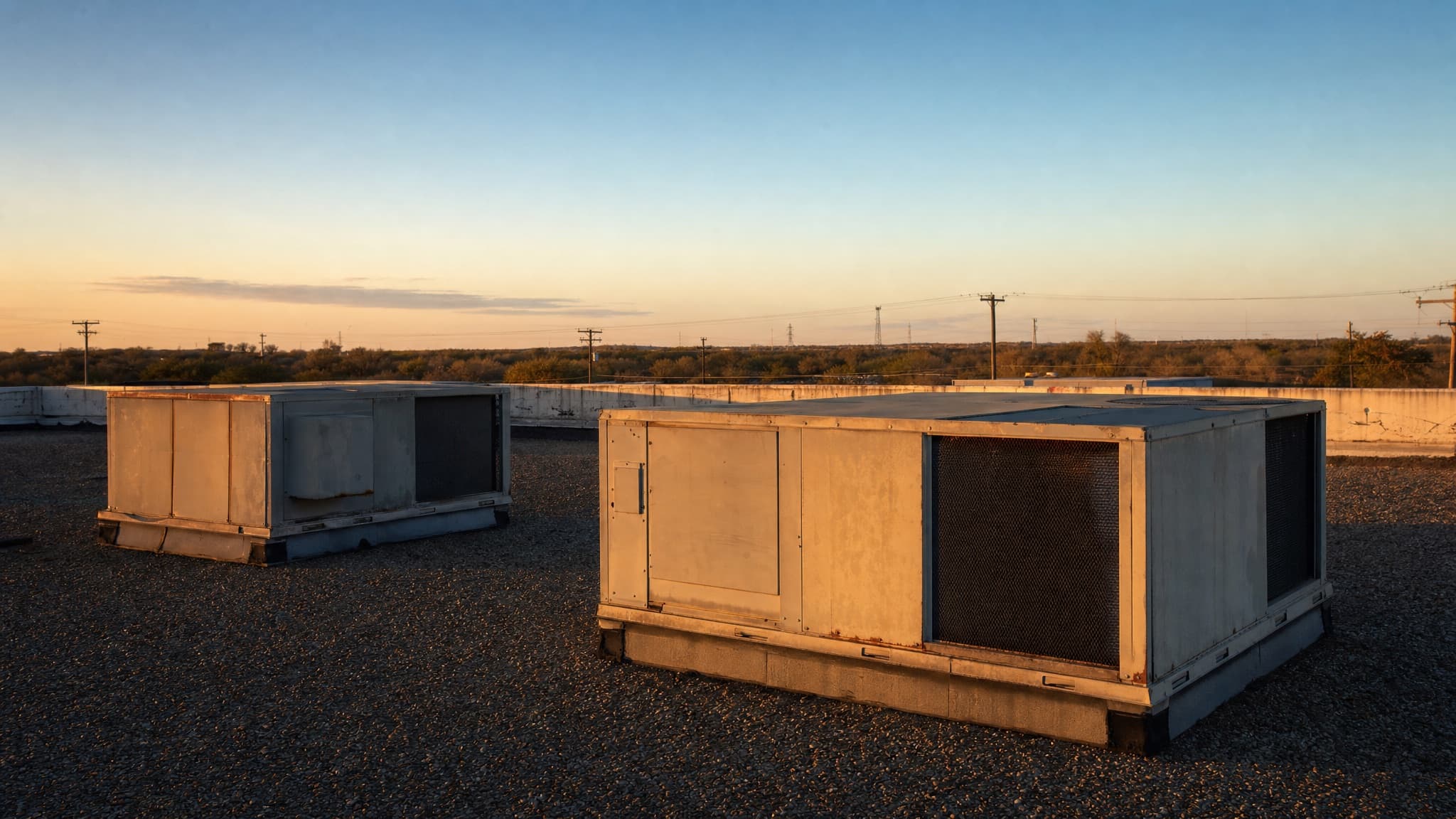 Two weathered rooftop package units on the gravel-ballasted roof of a low-slope commercial building at sunset, with a flat treeline and utility poles on the horizon.