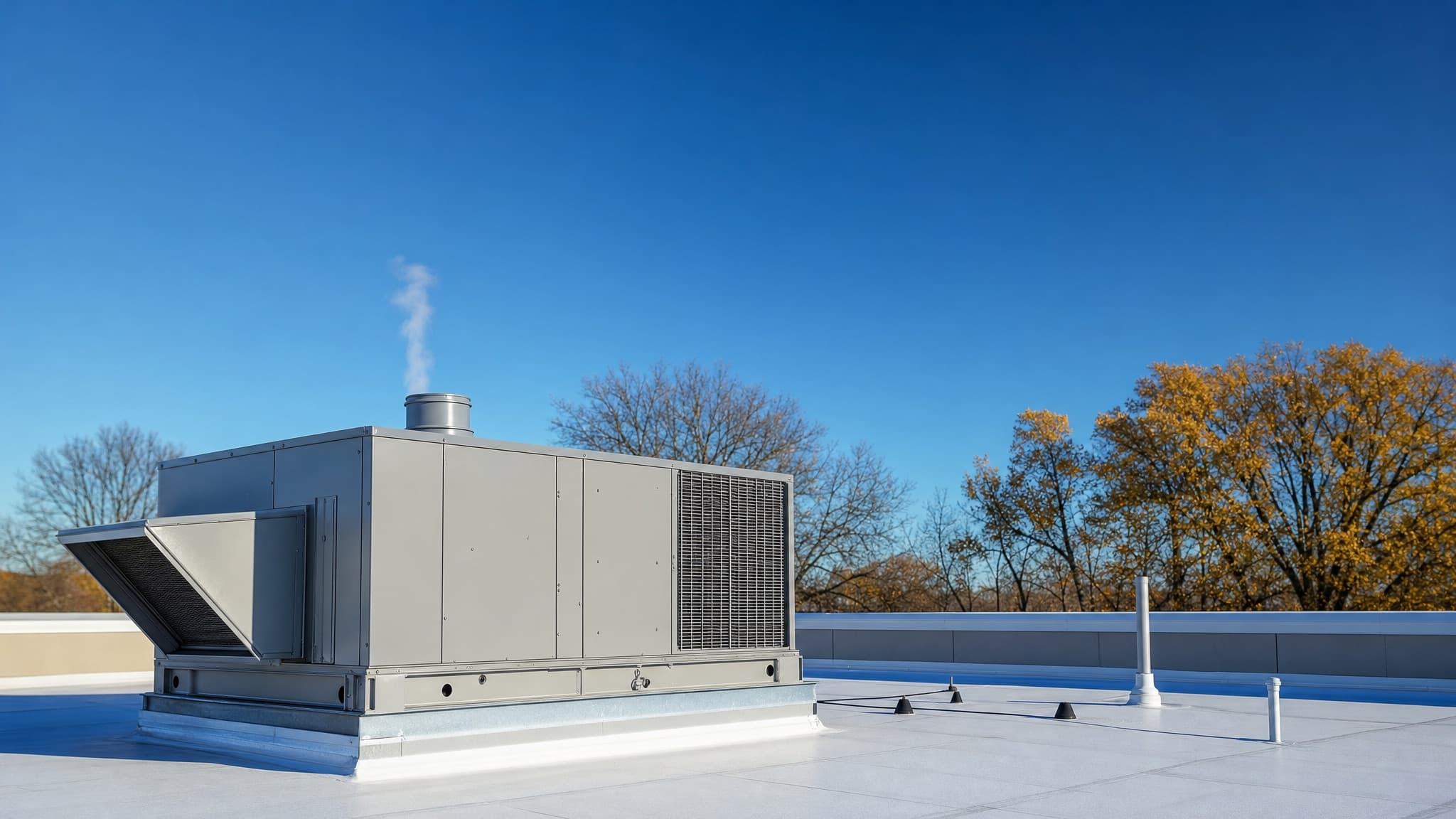 A modern rooftop HVAC unit on a white commercial roof on a clear autumn morning, with a wisp of vapor rising from its vent and gold-leaved trees beyond the roof edge.