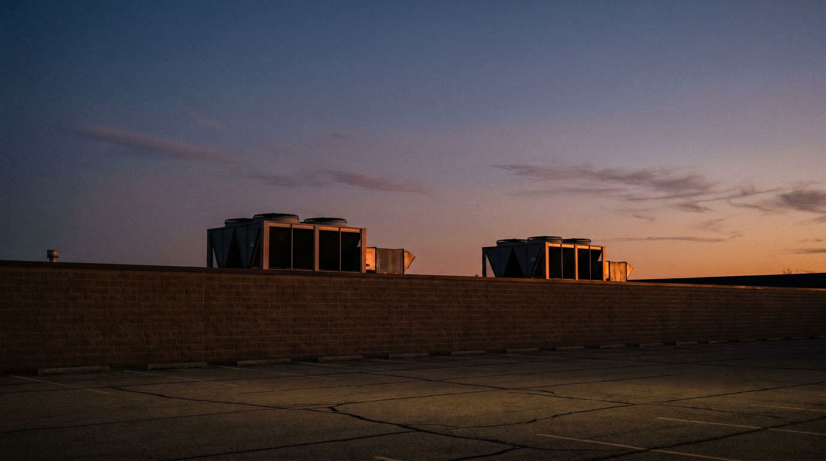 Low-angle view of a commercial building's brick parapet with rooftop HVAC units silhouetted above against a dusk sky. The landlord-tenant boundary made visual.